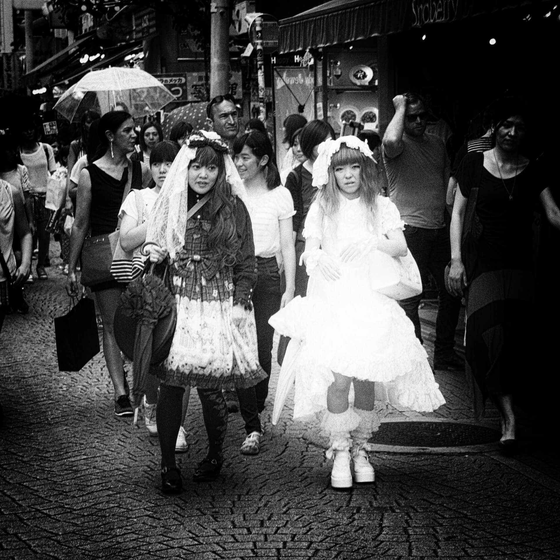 Two women dressed in elaborate lolita fashion walking through a bustling street market, captured in a high-contrast black and white photo.