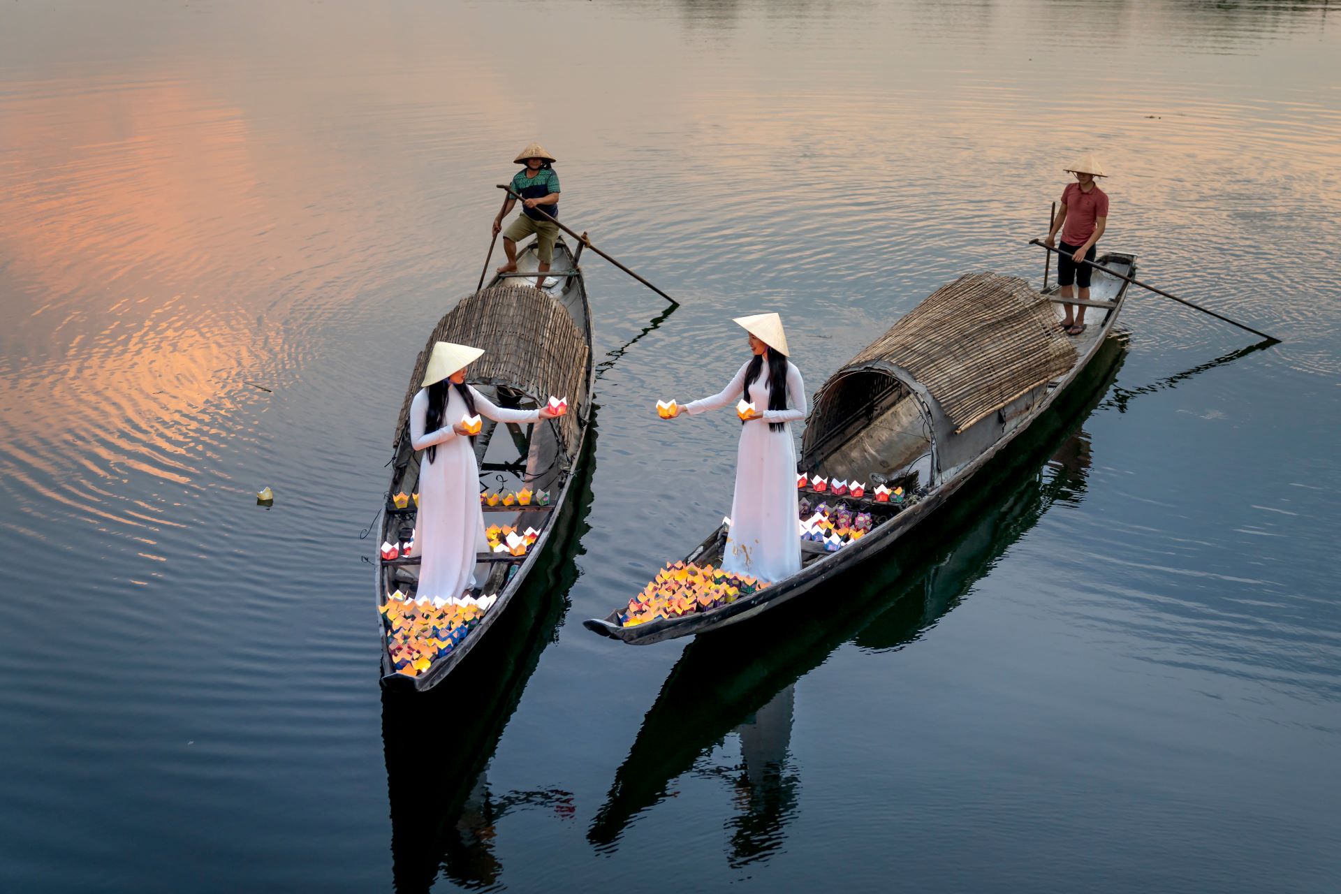 Three women in traditional vietnamese dresses stand on wooden boats, selling colorful flowers on a calm river at dusk.